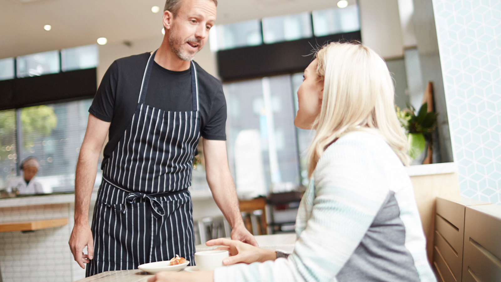waiter-customer-talking-1600x900 A man talking to a woman seated at a table in a café.